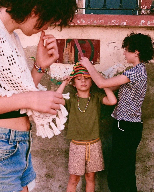 Three children in front of a weathered wall. One child is being helped with a colorful hat, while another looks on with curiosity.