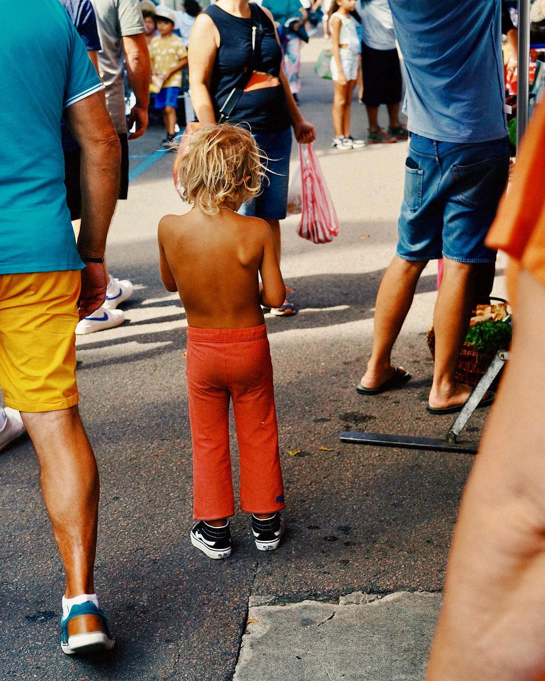 Child stands on pavement in a crowd wearing orange pants and black-and-white shoes; people are on either side and in the background.