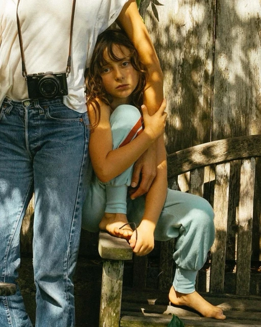 Young girl sits on a wooden bench next to an adult with a camera outdoors, with a weathered wooden fence in the background on a sunny day.