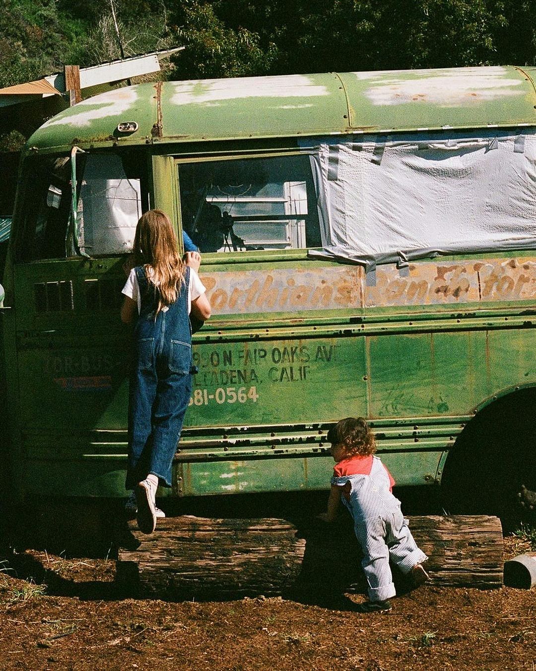 Two children in overalls play on a vintage green bus in an outdoor, sunny rural setting, next to logs and foliage.
