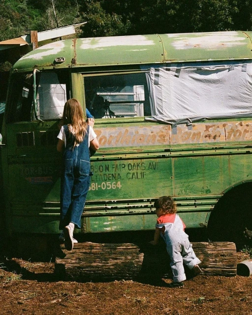 Two children in overalls play on a vintage green bus in an outdoor, sunny rural setting, next to logs and foliage.