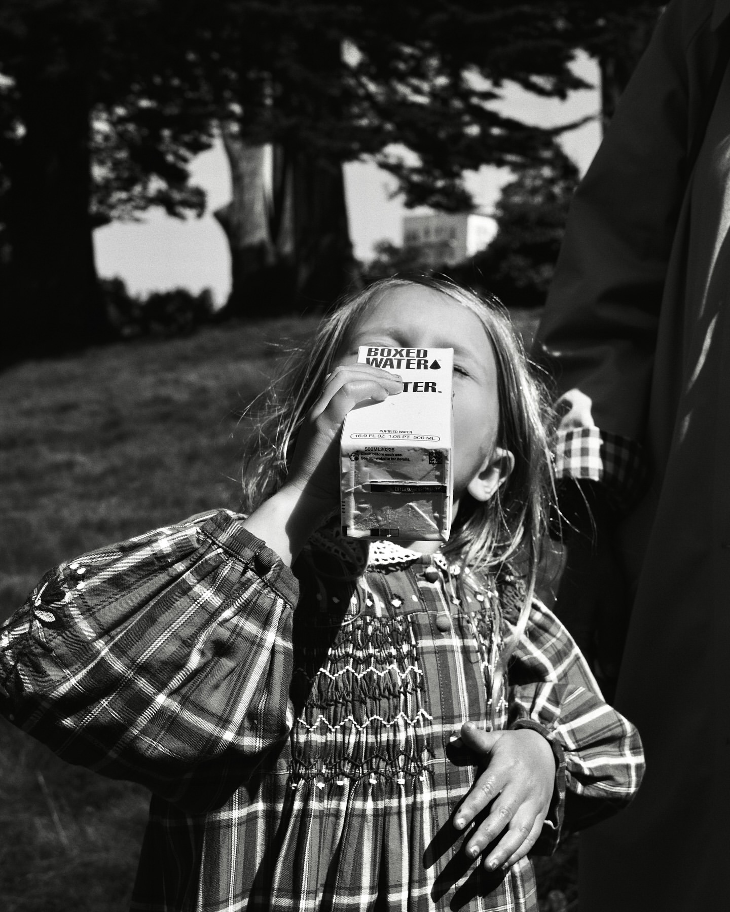A child with blonde hair drinks from a Boxed Water carton outside on a grassy lawn in a black and white photograph.