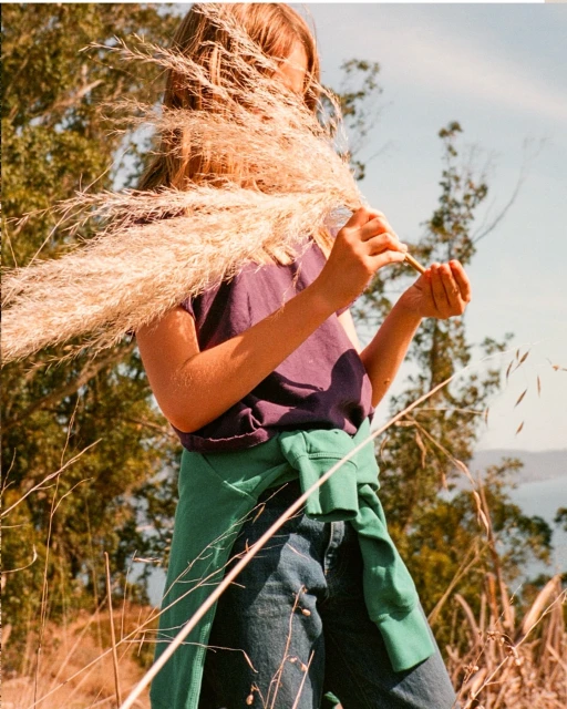 Girl stands on a grassy hill partially obscured by pampas grass with a tied green sweater. The background reveals a blue sky and body of water.