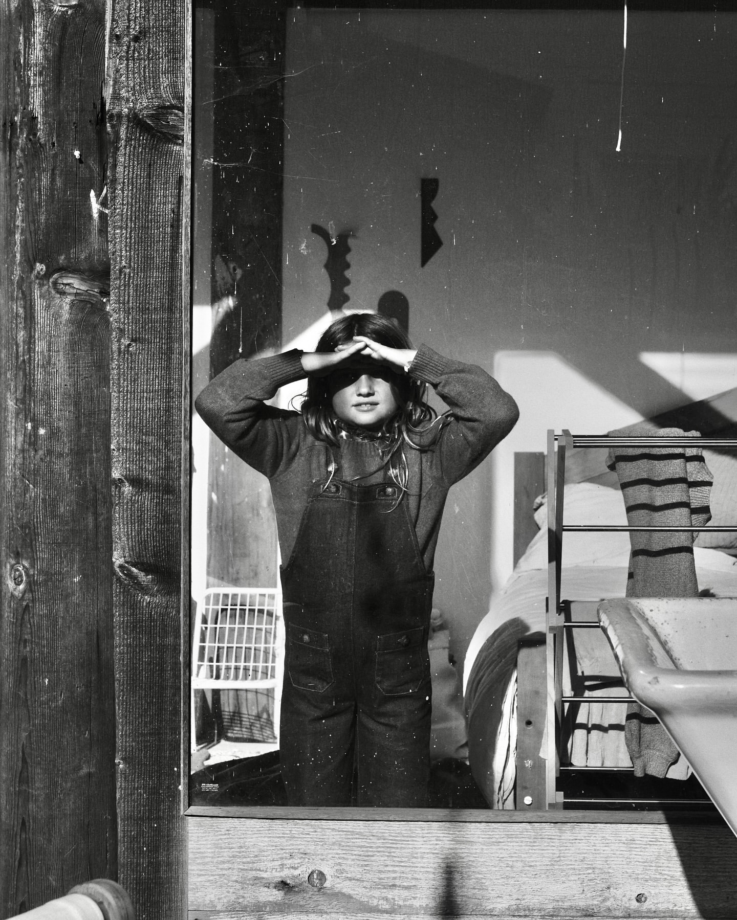 Black and white portrait of a young girl in overalls standing indoors, looking directly at the camera with her hands on her forehead.