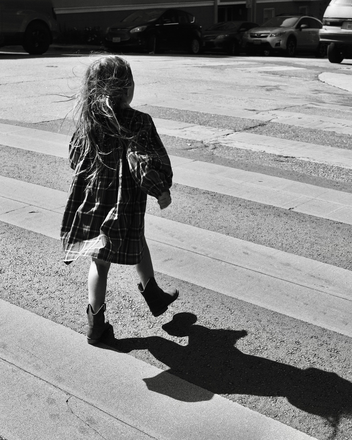 Young child with long hair walks across a crosswalk wearing a plaid dress and cowboy boots, casting a long shadow on the road.