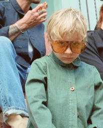 Young child with blonde hair wearing amber-tinted sunglasses and a green button-down shirt, captured by Claire Guarry.