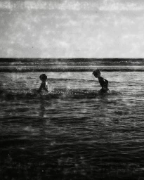 Two children splashing in the ocean waves under an overcast sky captured in black and white photography by Claire Guarry.