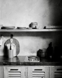 Monochrome kitchen interior featuring wooden cabinets, a stone countertop, a shelf with stacked ceramic plates and bowls, and wooden cutting boards