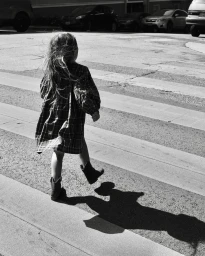 A young child walking across a street in a plaid dress and boots, casting a long shadow, captured by Claire Guarry.