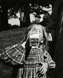 Young child in a plaid dress drinking from a boxed water carton outdoors, captured in black and white by Claire Guarry.