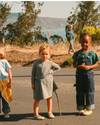 Four children outdoors near a body of water, wearing denim overalls and casual clothing, photographed by Claire Guarry.