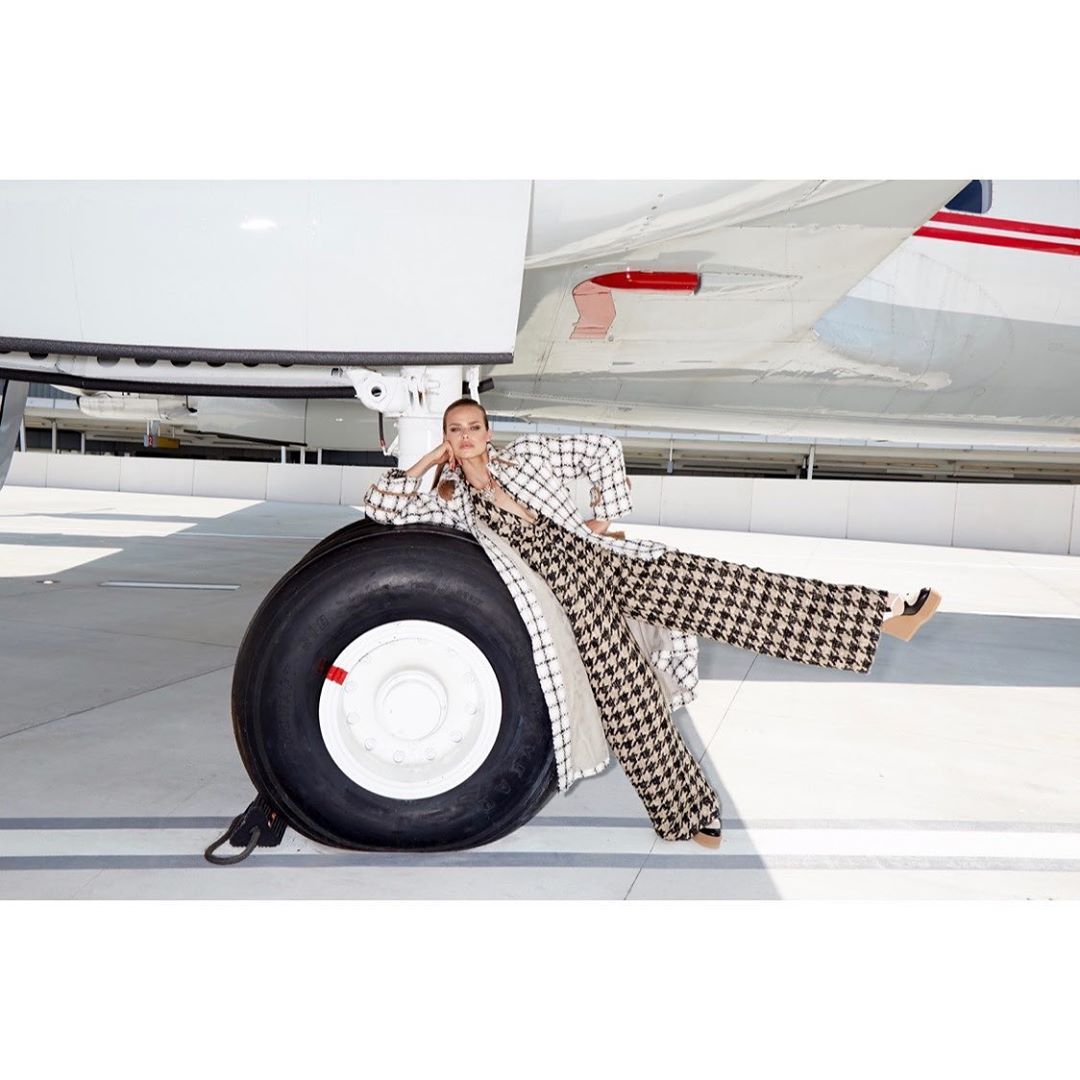 Woman in a houndstooth jumpsuit and jacket poses on an airplane wheel on the tarmac, looking at the camera.