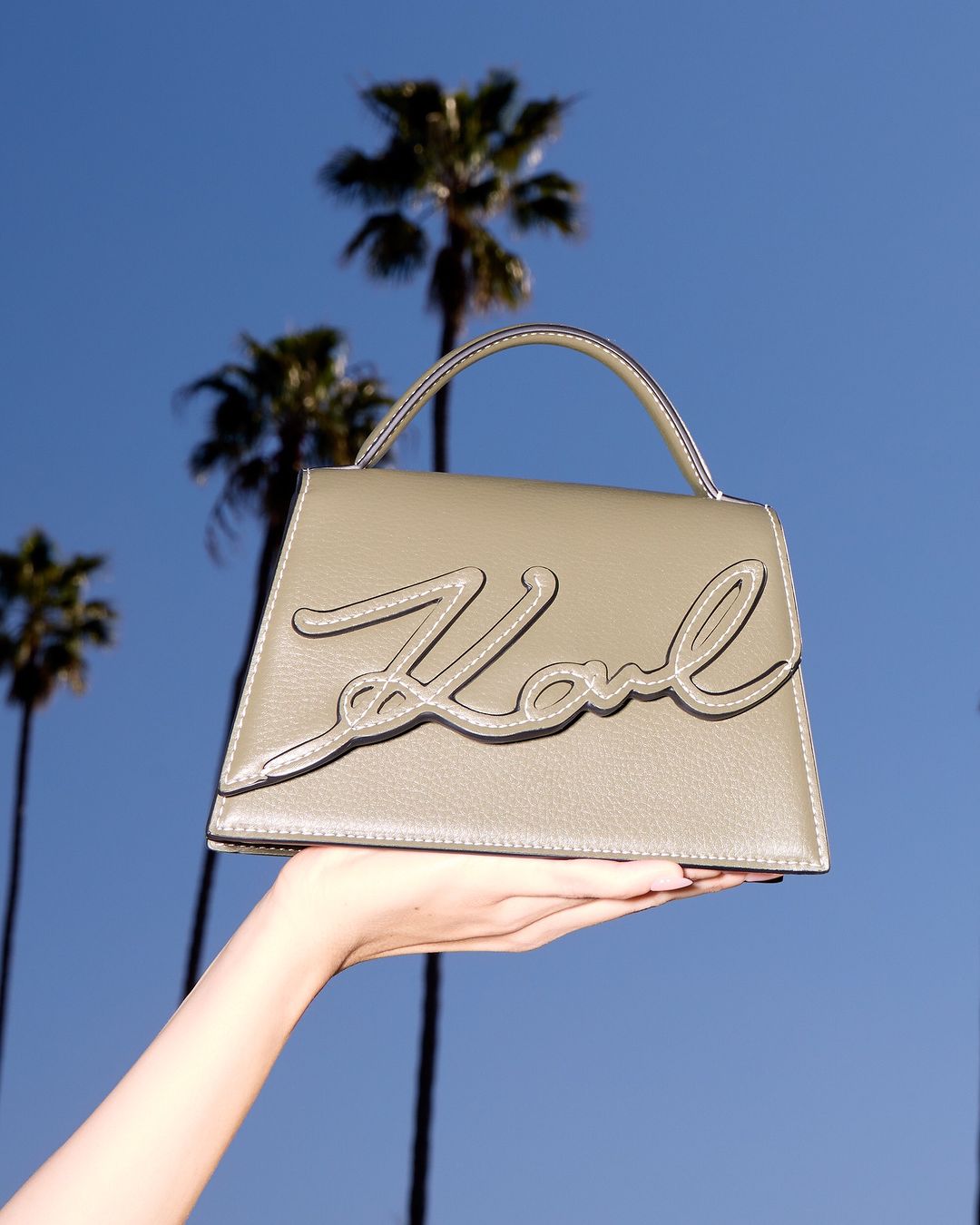 Hand holding a taupe leather handbag with cursive logo against a bright blue sky and tall palm tree silhouettes