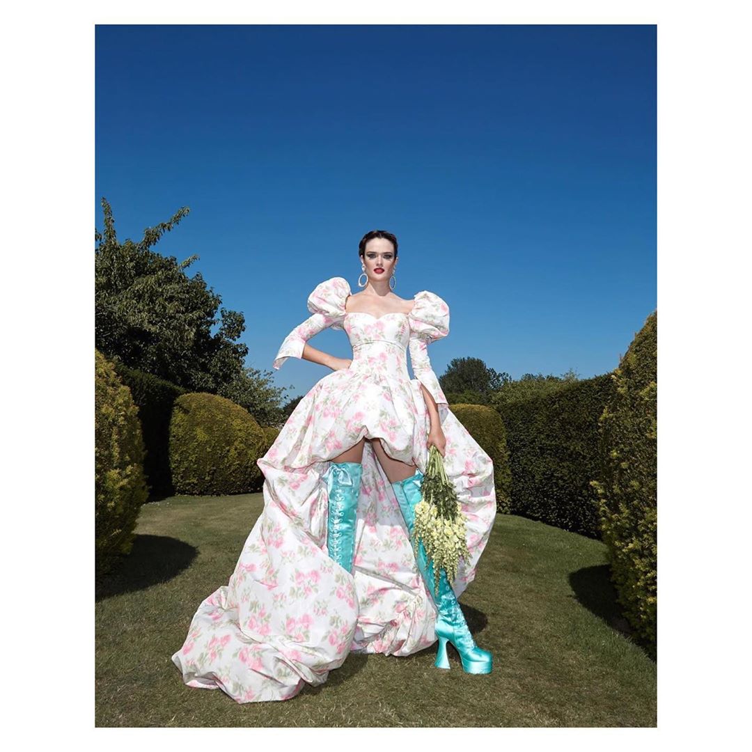 A woman in a floral puff-sleeve dress and blue platform boots poses in a garden against a blue sky. She holds a bouquet of white flowers.