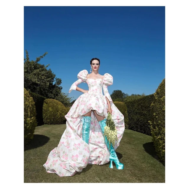 A woman in a floral puff-sleeve dress and blue platform boots poses in a garden against a blue sky. She holds a bouquet of white flowers.