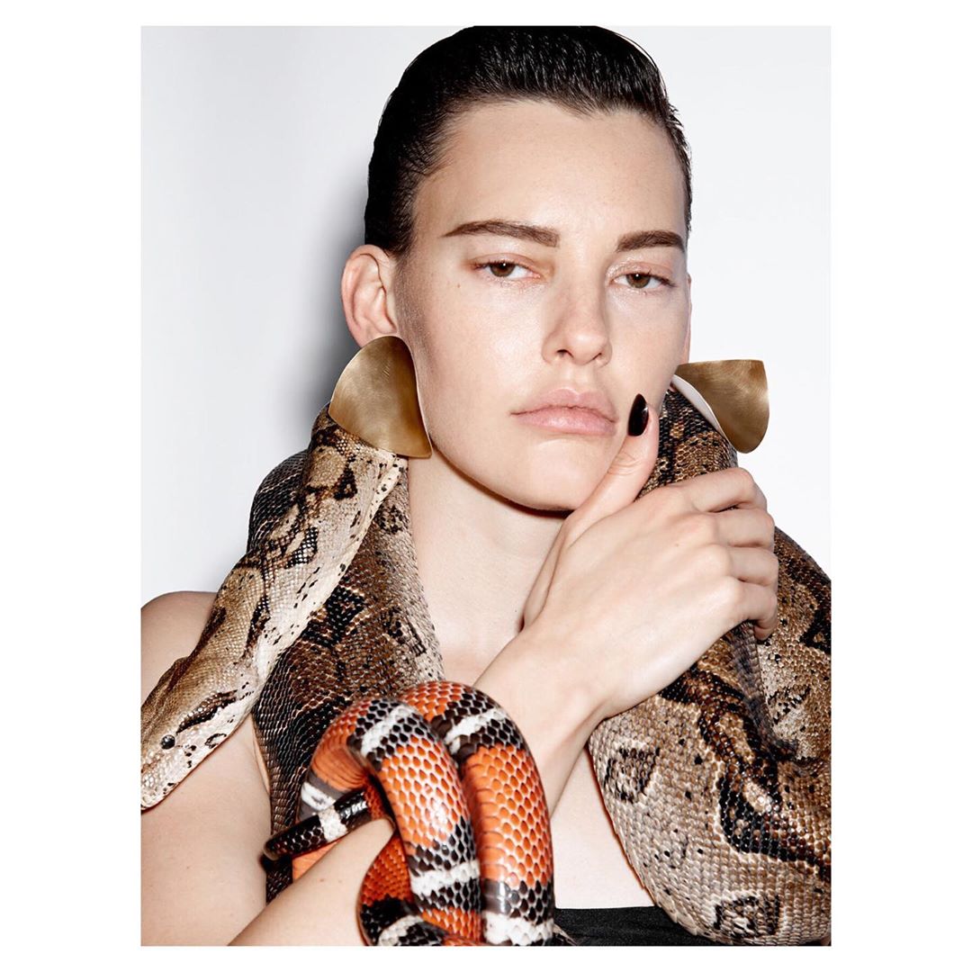 Headshot of a woman with dark hair, wearing large gold earrings and posing with two snakes around her neck against a white background.