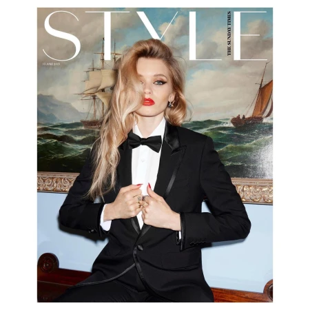 Woman wearing a black tuxedo and bowtie with blonde hair standing against a maritime painting