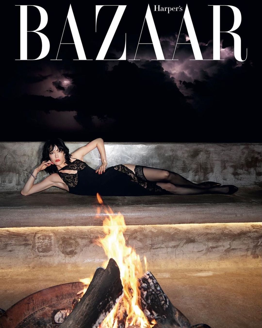 Fashion editorial image featuring a woman in a black dress lying on a concrete platform near a campfire, with the Harper's Bazaar logo above.