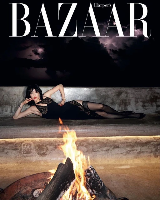 Fashion editorial image featuring a woman in a black dress lying on a concrete platform near a campfire, with the Harper's Bazaar logo above.