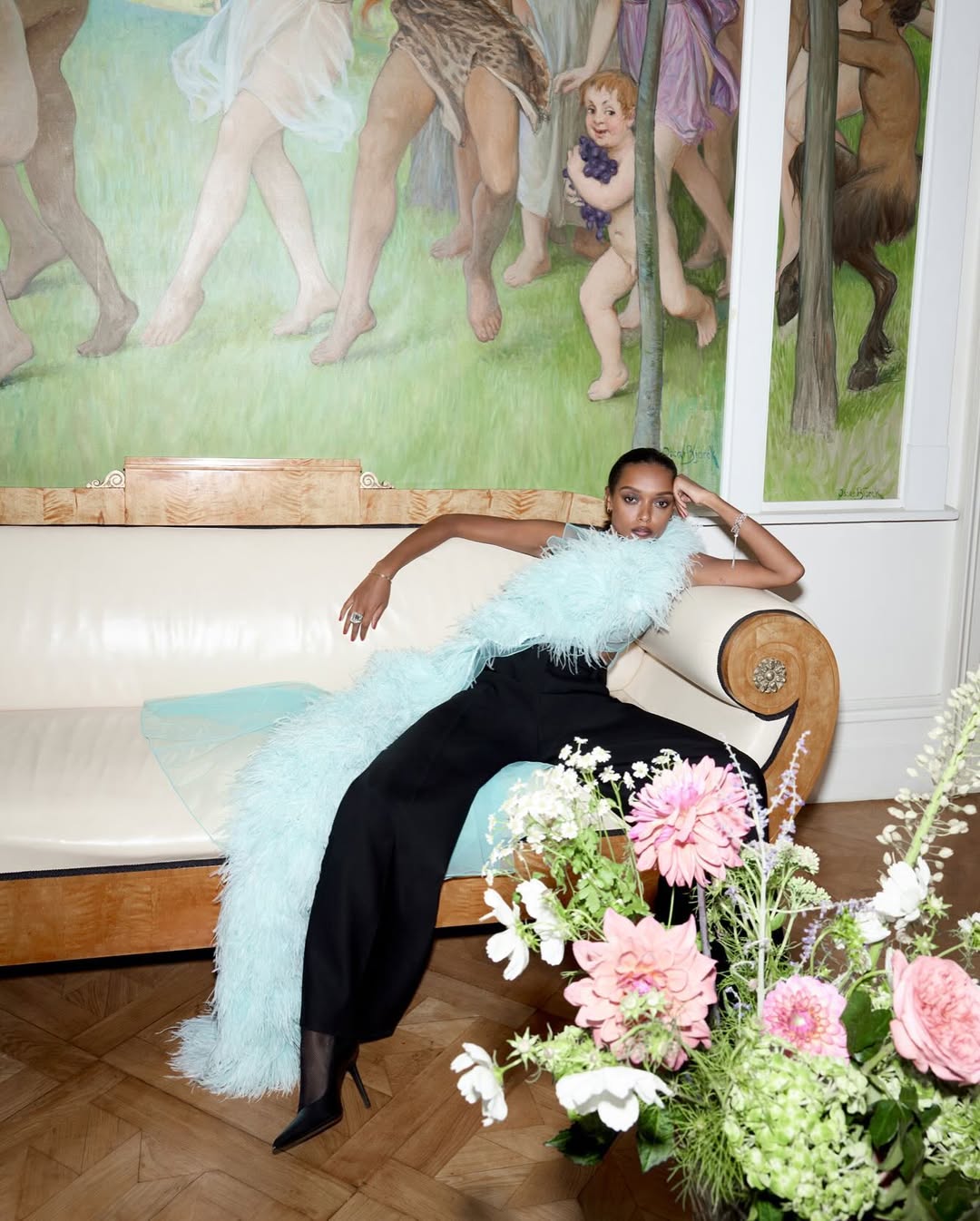 Elegant woman in black jumpsuit poses on a cream sofa with a large floral arrangement in a room featuring an artistic mural.