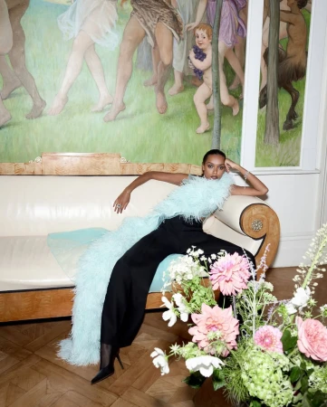 Elegant woman in black jumpsuit poses on a cream sofa with a large floral arrangement in a room featuring an artistic mural.