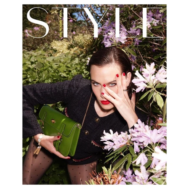 Fashion magazine cover featuring a woman in a dark jacket with a green purse, floral background, and bold red lips and nail polish.