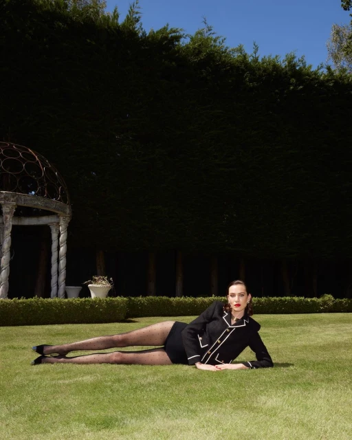 Elegant woman in black jacket and shorts lies on green lawn against a green hedge and blue sky in outdoor fashion setting.