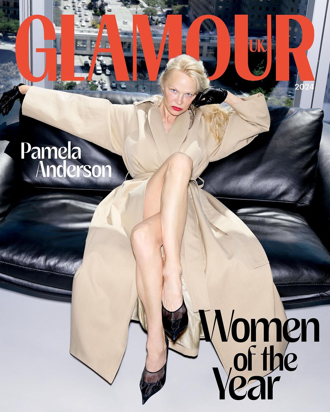 Pamela Anderson poses on a black leather couch for Glamour UK's 2024 Women of the Year issue, set against a cityscape backdrop.