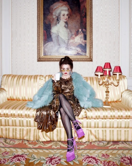 Woman in a gold dress and blue faux-fur coat sits on a striped sofa, holding a teacup with an ornate painting in the background.
