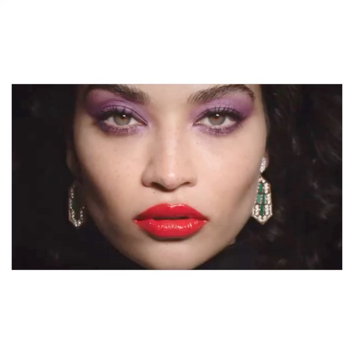 Close-up of a woman's face with vibrant purple eyeshadow and glossy red lipstick against a dark background, showcasing beauty and makeup.