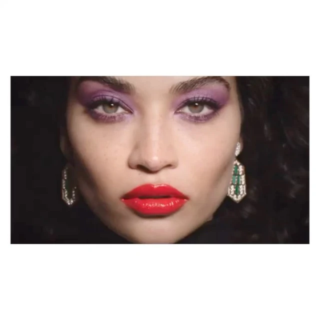 Close-up of a woman's face with vibrant purple eyeshadow and glossy red lipstick against a dark background, showcasing beauty and makeup.