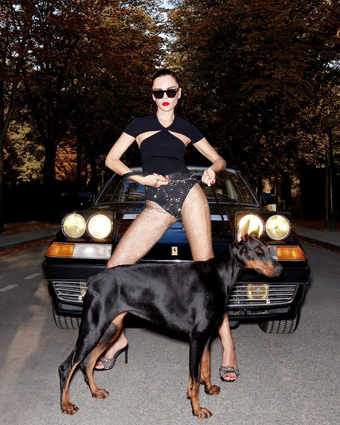 Woman poses with a black Doberman dog and a black Ferrari on an avenue lined with trees in fall foliage during daylight.