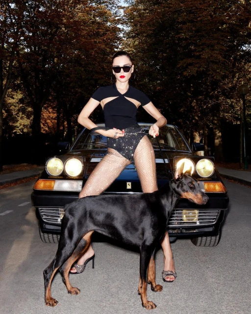 Woman poses with a black Doberman dog and a black Ferrari on an avenue lined with trees in fall foliage during daylight.