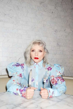 Debbie Harry in a light blue floral blouse holding a knife and fork at a marble table, photographed by Claire Rothstein.