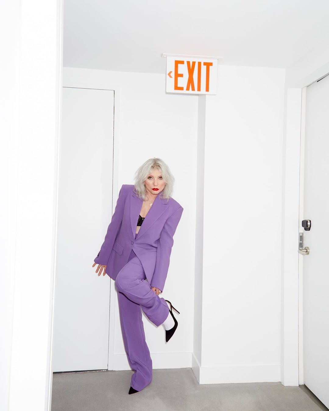 Adult woman in a lavender suit poses in a white hallway with an orange 'Exit' sign above. She is wearing black pumps and white socks.