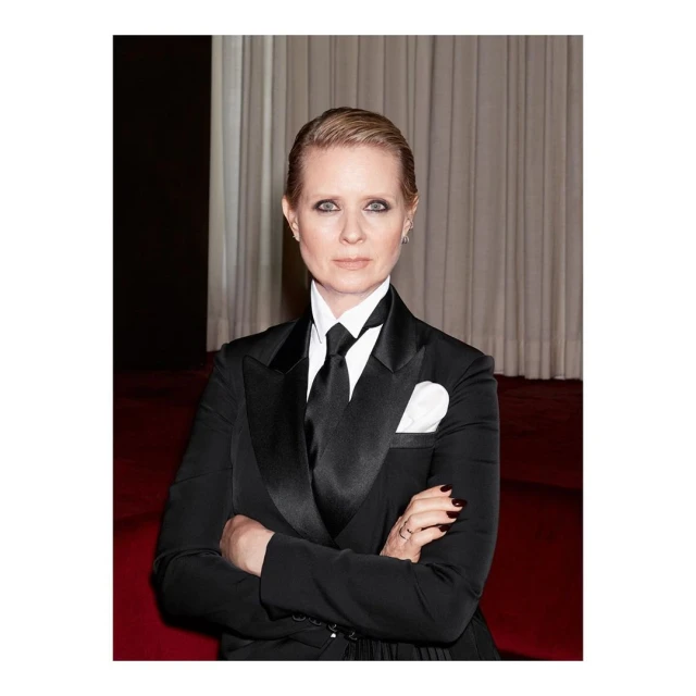 Stylish portrait of a woman in a black suit, crisp white shirt, and a black tie, standing indoors against a soft-lit backdrop.