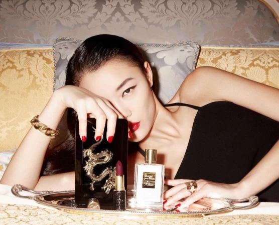 Asian woman laying on bed with red lipstick, black dress, and gold jewelry. On a silver tray are perfume, lipstick, and a gold dragon.