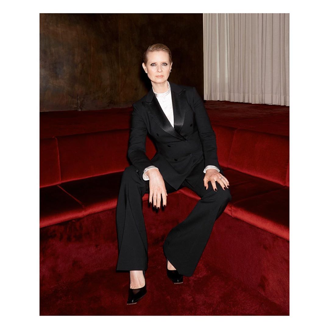 Cynthia Nixon sits on a red velvet couch wearing a black tuxedo and black patent leather heels in an indoor environment.