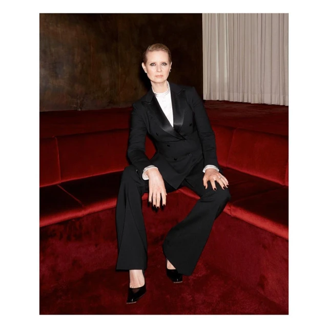 Cynthia Nixon sits on a red velvet couch wearing a black tuxedo and black patent leather heels in an indoor environment.