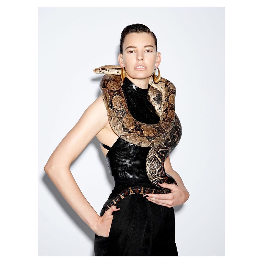 Fashionable adult female poses in a studio with an exotic snake wrapped around her shoulders and torso, wearing a black dress.