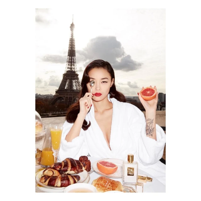 Woman in robe poses at breakfast in Paris with Eiffel Tower in background. Croissants, juice, and perfume are on the table.
