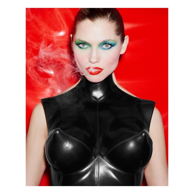 Glamour shot of a female model with bold makeup in a black latex top, set against a vivid red backdrop with wisps of smoke.