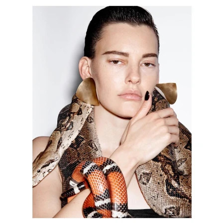 Portrait of a woman holding a snake draped over her shoulders wearing large triangular gold earrings against a white background