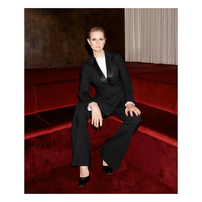 Cynthia Nixon seated in a black double-breasted suit on a deep red banquette in Paris, photographed by Claire Rothstein.