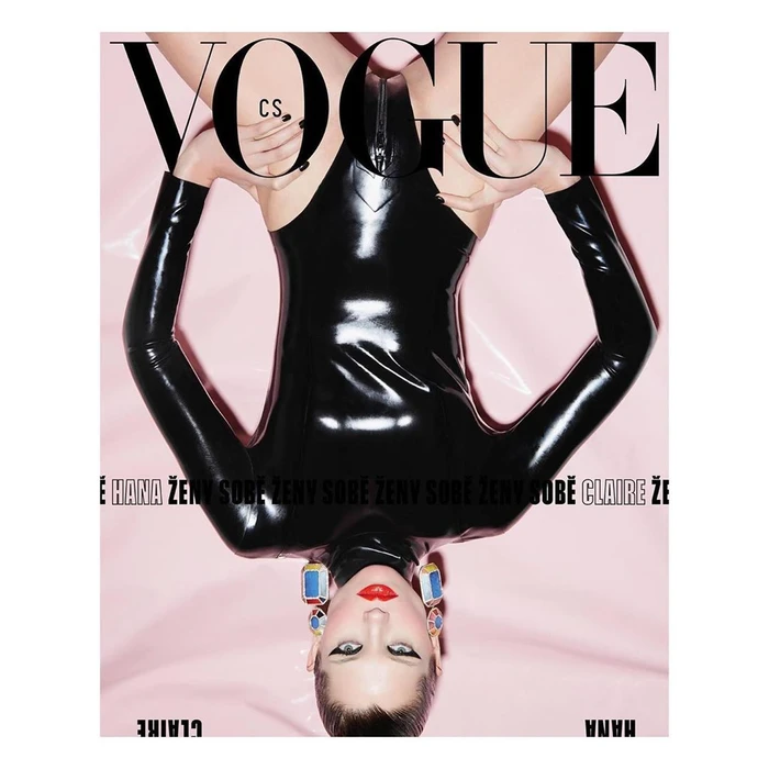 Upside-down model wearing a glossy black latex bodysuit and large colorful earrings against a pale pink background