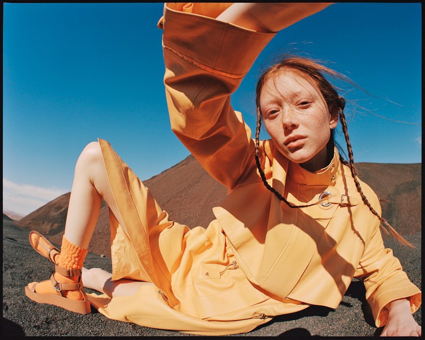 A red-haired woman in a yellow outfit poses while lying on a dirt mound against a blue sky in an outdoor fashion shot.