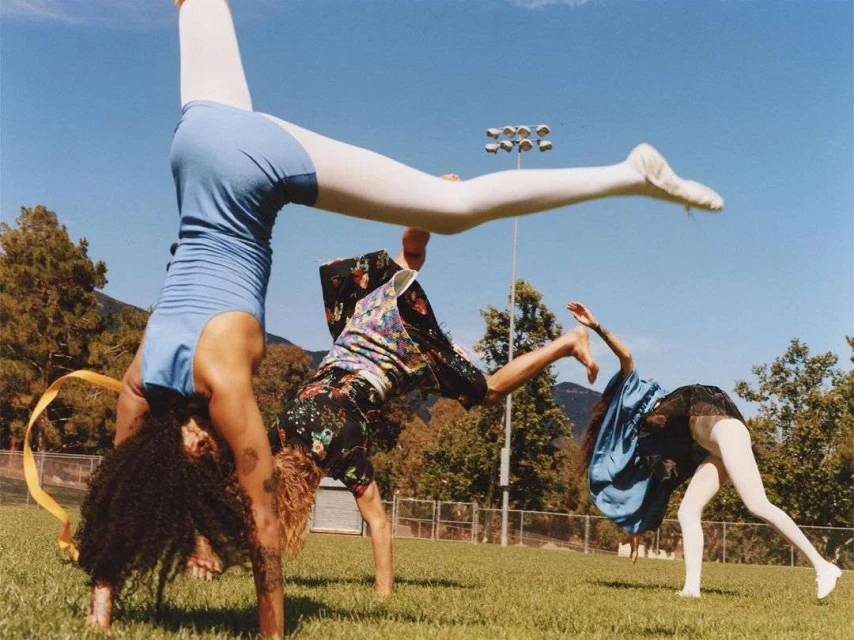 Three people doing cartwheels in a grassy field on a sunny day, wearing athletic attire. Mountains are visible in the background.