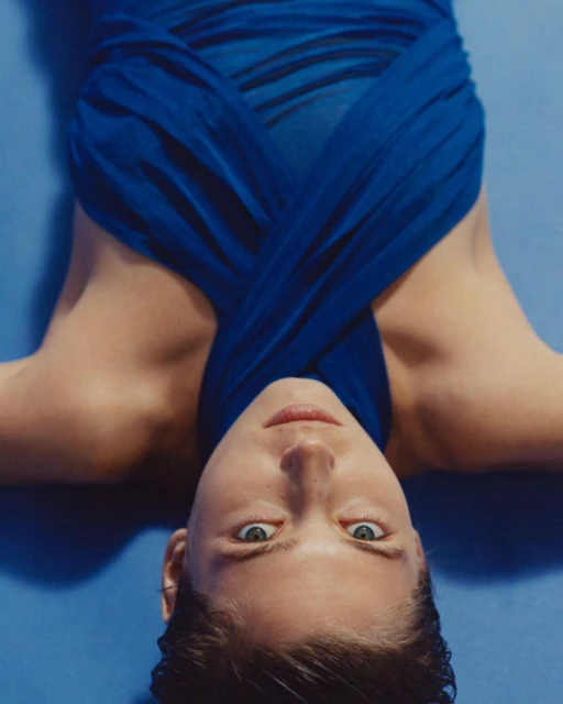Eye-level portrait of an adult woman with a blue garment, lying on a light blue background, looking directly at the camera.