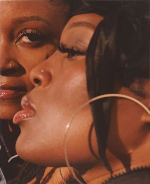 Close-up portrait of two dark-skinned adult females with nose piercings, glossy lips, and hoop earrings in a dimly lit indoor setting.