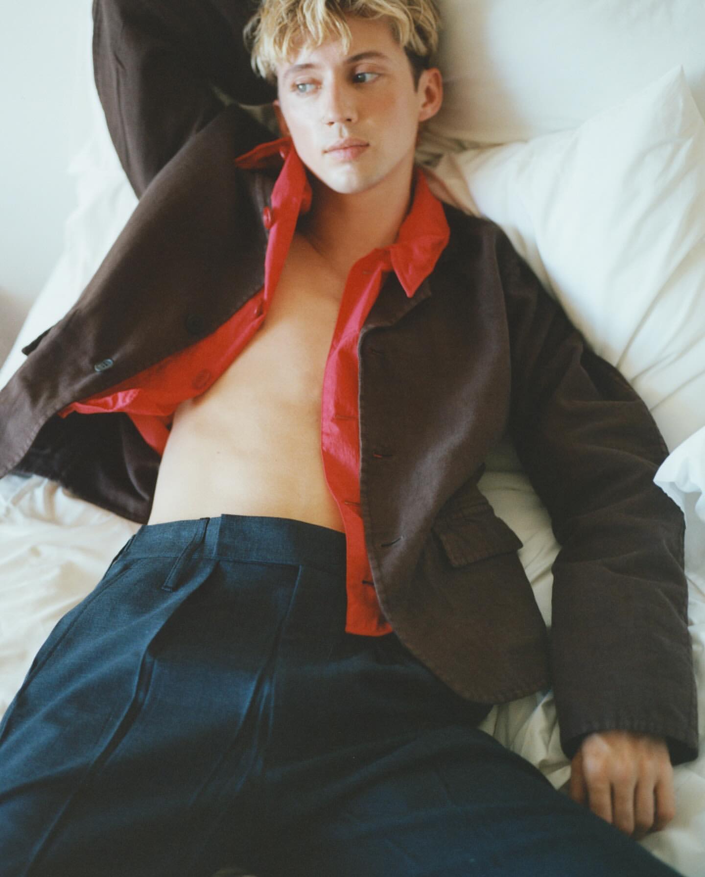 Young adult with blond hair poses in bed, wearing a brown jacket and blue pants, showcasing fashion and lifestyle elements.